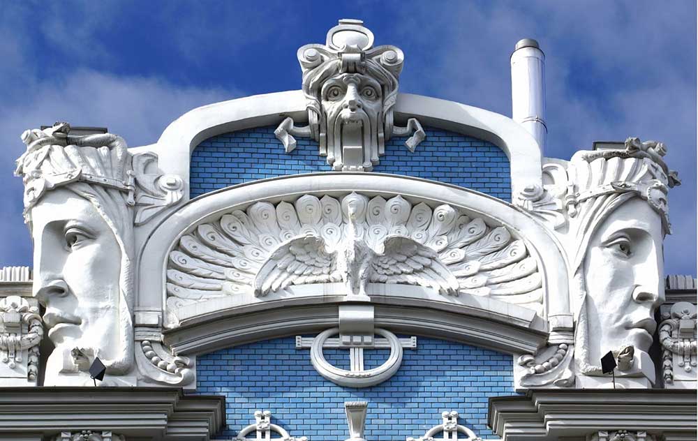 Best Riga 3 Days Itinerary: Exploring ‘Paris of the North’ 19 The top of a building in the Art Nouvea district in Riga. The tiles are blue and there are two large faces lookin in opposite directions. There is another face at the top centre, and a peacock in the midle centre.