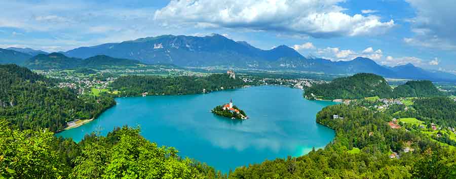 An aerial view of Lake Bled from Ojstrica showcases a small islet at the center, surrounded by forest trees, with a breathtaking view of the mountains in the distance.