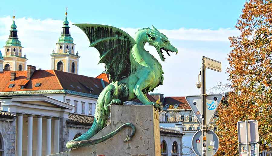 A green dragon statue stands on the Dragon Bridge in Ljubljana, with houses in the background.