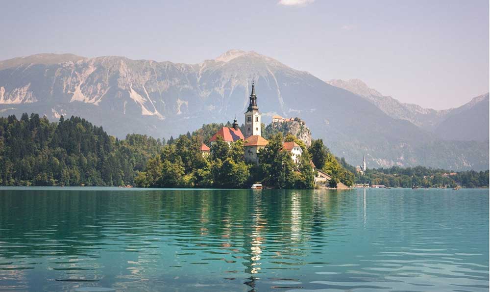 Lake Bled features a glacial lake with a small islet that houses a charming church adorned with a red-tiled roof and a baroque bell tower, all set against a backdrop of majestic mountains.