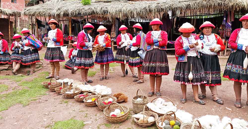 G Adventures Inca Trail Review: What to Expect on The Inca Trail Trek to Machu Picchu 13 Women at the Weaving Cooperative which is supported by G Adventures, they are wearing traditional peruvian clothing - a bowl shaped red and white hat, a red cardigan over a white embroidered shirt and a knee-length blak skirt with embroudery around it.
