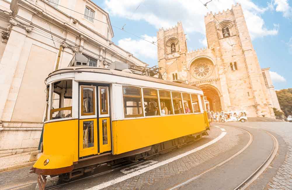 Lisbon Cathedral features sandstone walls with a carved flower at the center, and a yellow tram passing by near the cathedral.