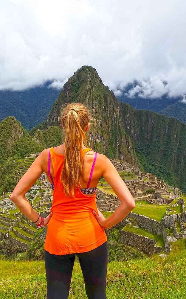 G Adventures Inca Trail Review: What to Expect on The Inca Trail Trek to Machu Picchu 21 A photo of a woman in an orange tshirt (me) standing in front of Machu Picchu there are white clouds in the background.