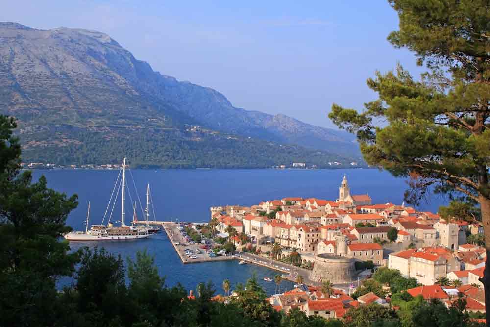 the city of korcula in croatia with blue sea and a mountain behind it, there are some boats in the sea surronding the city