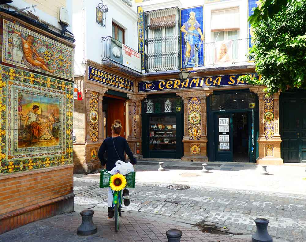 A woman seen from behind, riding a bicycle with a green crate containing a large sunflower, navigates a cobblestone square. She is approaching 'CERAMICA SANANA', a traditional shop with an intricate blue and yellow facade. To the left, a large, ornate tile mural adorns the side of another building, all set under the warm sun of the Streets of Triana district.