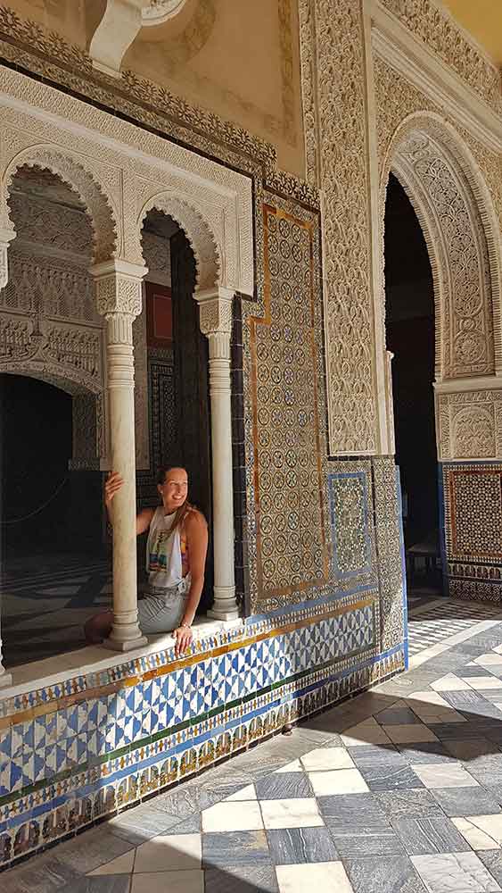 A woman (me) in a white graphic tank top and light wash denim overalls leans against a detailed white carved column, smiling in a room decorated with blue, yellow, and black geometric-patterned tile and intricately-carved walls.