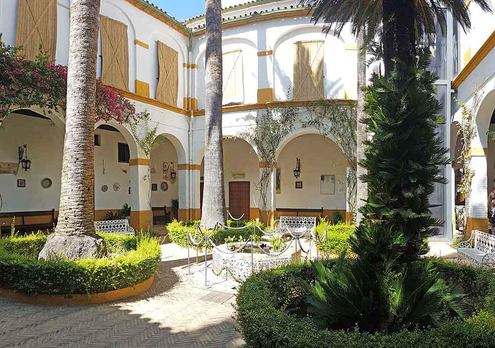A sun-drenched traditional Andalusian courtyard in Seville, featuring white-arched cloisters with yellow trim and towering palm trees. Lush green hedges and white wrought-iron benches surround a central tiled fountain, making this picturesque patio a quintessential addition to any Spain bucket list.