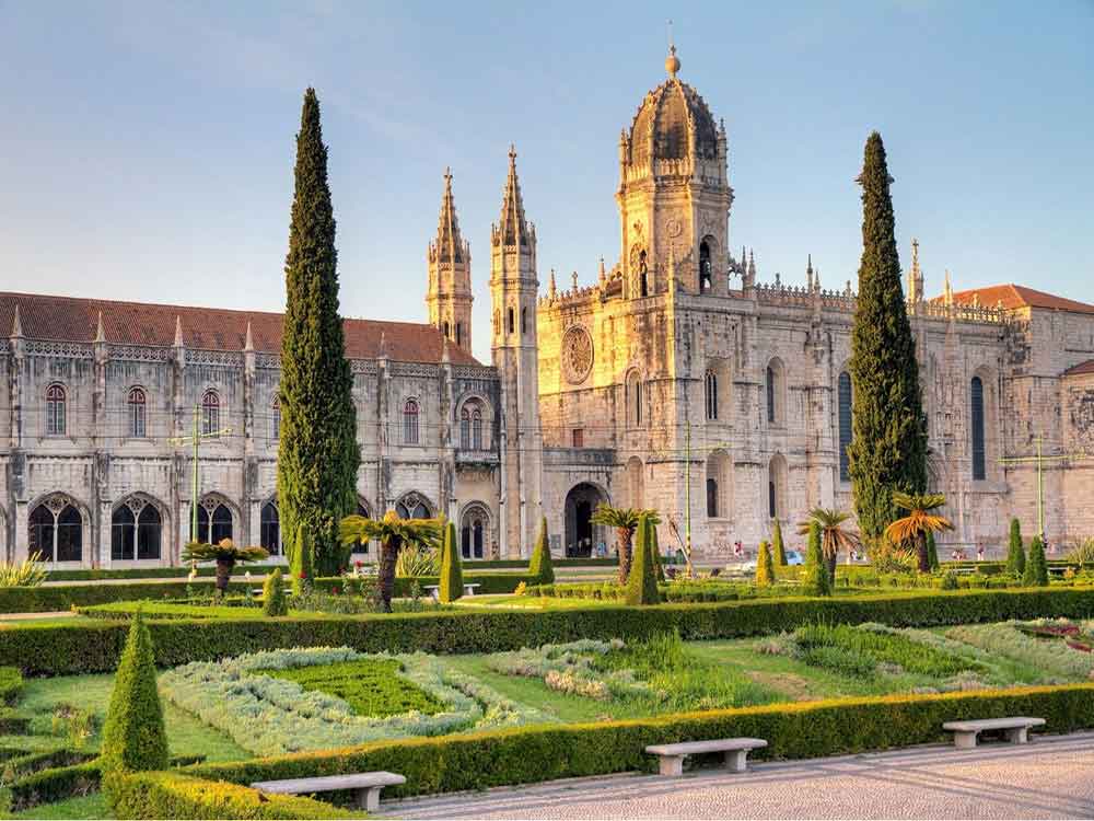 Jeronimos Monastery in Lisbon, Portugal, showcases its towering turrets and spires with a well-manicured garden at the front during sunrise.