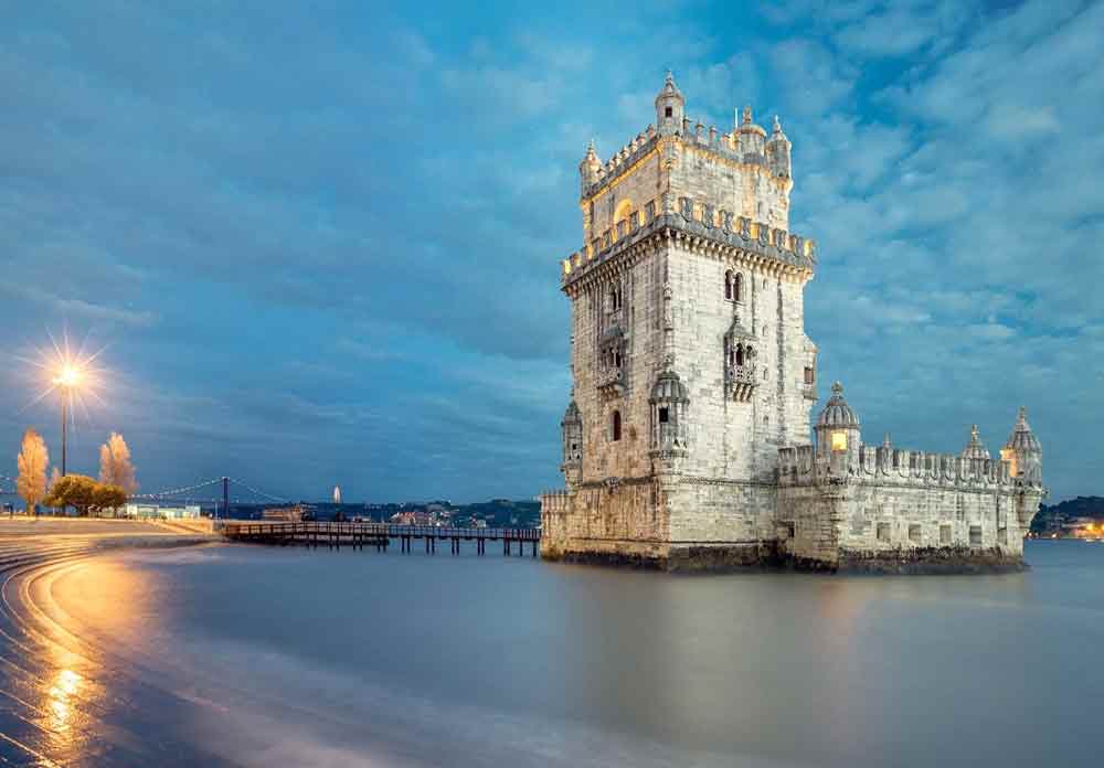 Belem Tower in Lisbon stands on an island, featuring grayish-white watchtowers, battlements, and fortified walls, illuminated on an evening night.
