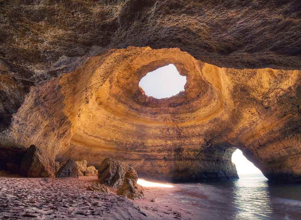 Benagil Cave in Algarve with an open-top cave sending a shaft of golden light into the sand with sandstone walls