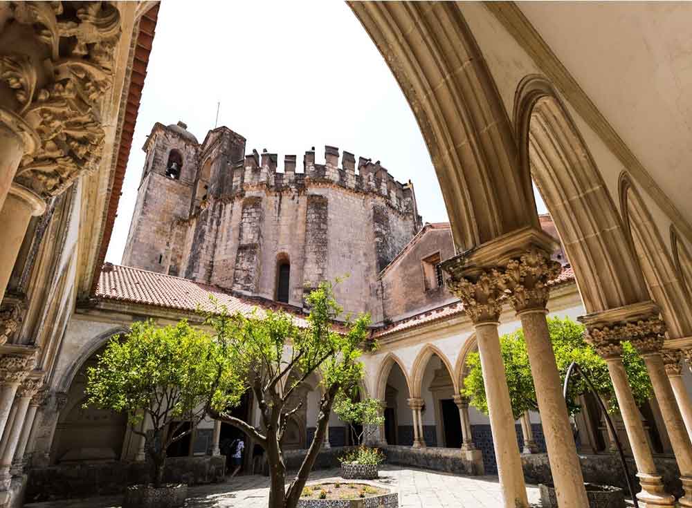 The Convent of Christ, a historic monastery located in Tomar, features archway hallways on brown stone with intricate details, with trees at the center.