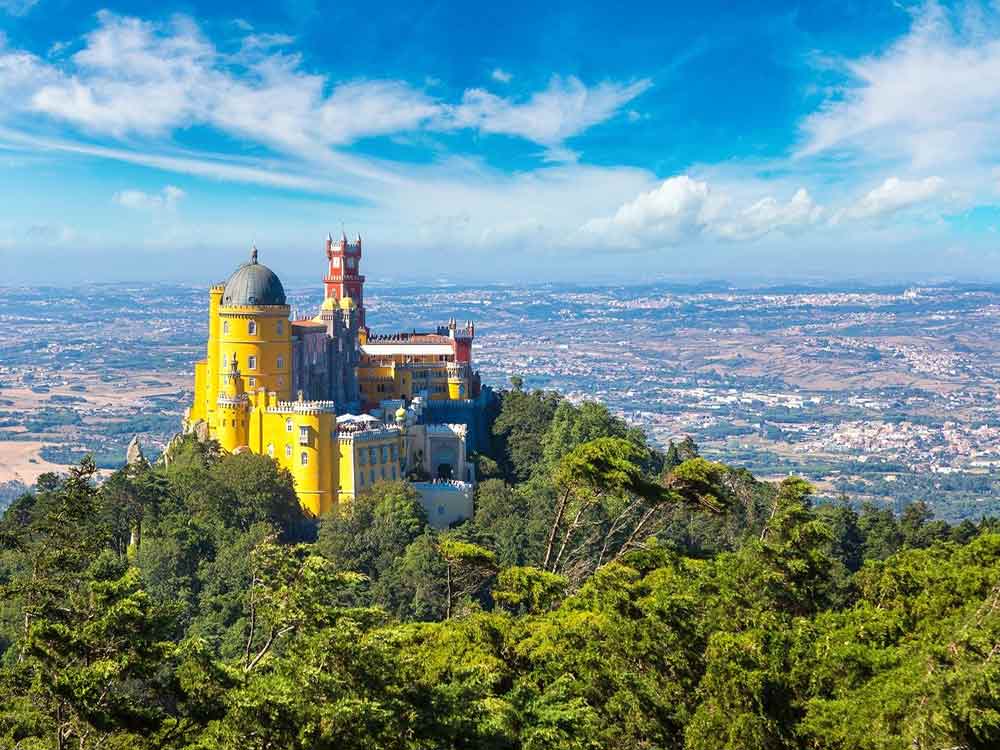 
Pena Palace in Portugal is painted with yellow, blue, and red walls, perched on top of a hill surrounded by lush trees, offering a stunning view of the landscape.