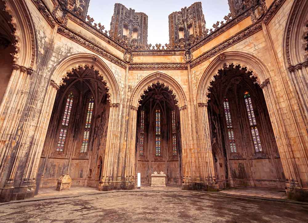 Batalha Monastery is an elegant, roofless Gothic-style architecture in a sandstone color, featuring multiple archways and ornate details.
