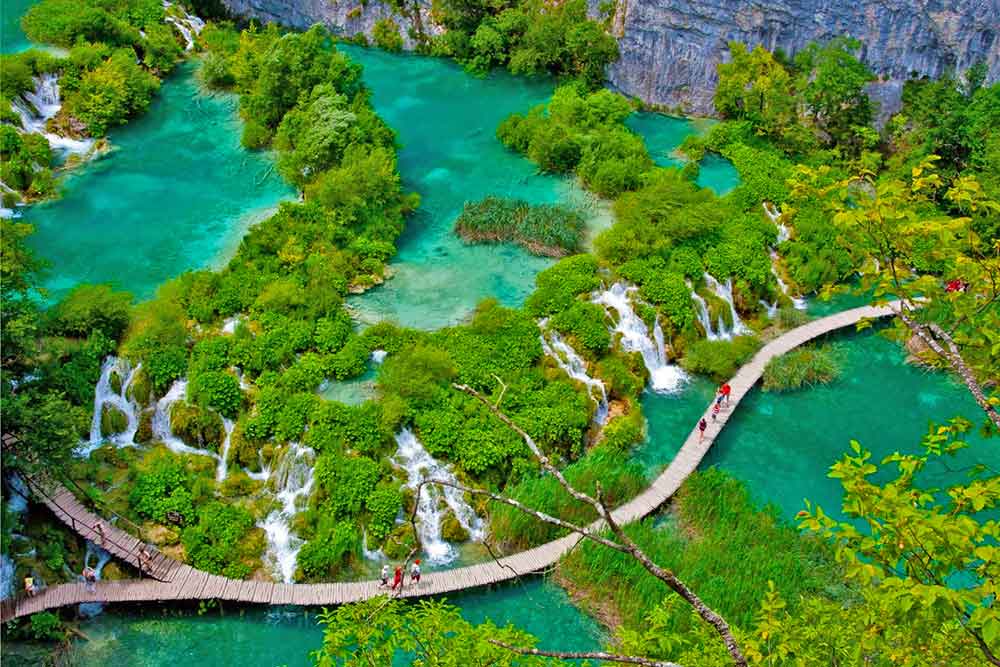 birds eye view of the Plitvice lakes in Croatia. Worthy of a adding to your Croatia bucktet list. A wooden walk way brdige meandering through waterfalls and trees. The trees are vibrant green and the water is turquoise blue.