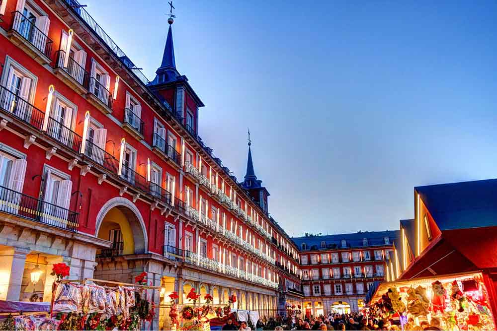 Madrid In Winter: Over 30 Things to Do In Winter in Madrid 2 Christmas Markets at Plaza Mayor with dusk sky in the background and yellow glow of the stalls