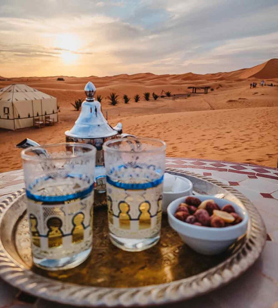 Two decorated glasses and a tea pot of mint tea with the sahara desert in the background.