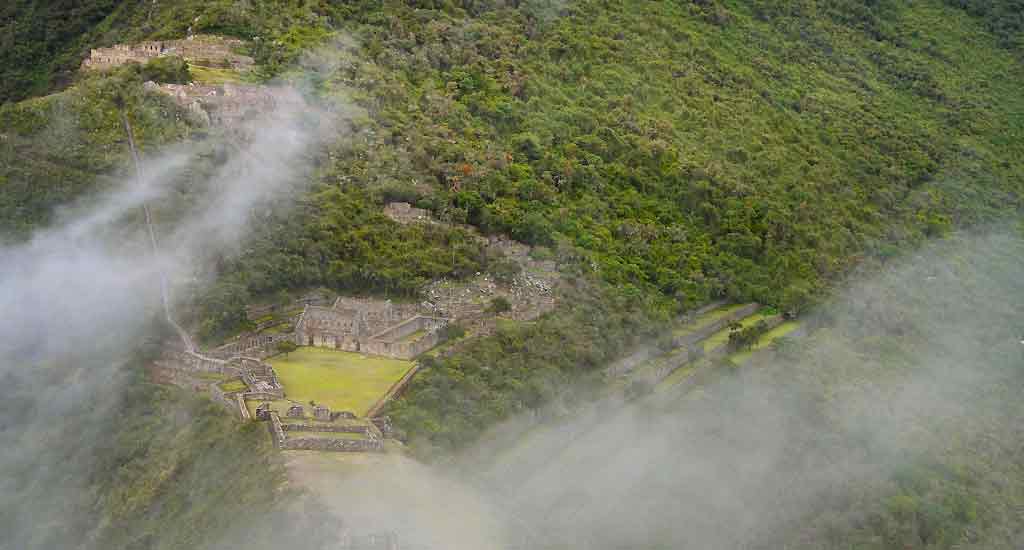 A large settlement of ruins at Choquequirao surrounded by trees on a grassy hillside and surrounded by clouds. 