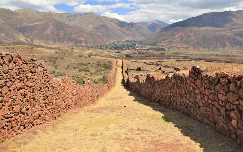 The Pikillacta Ruins in Peru showing a road between two dry stone walls with arid scenery and mountains in the background. 