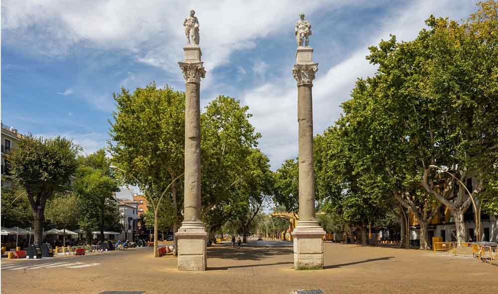 The historic Alameda de Hércules plaza in Seville, featuring two towering ancient Roman columns topped with statues of Hercules and Julius Caesar. The wide, tree-lined pedestrian promenade is paved with decorative tiles and surrounded by vibrant cafes, standing as one of the most iconic landmarks to visit in Seville.