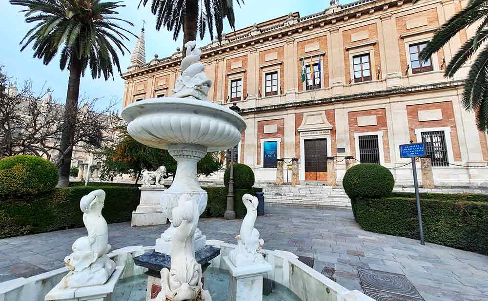 The grand facade of the Archive of the Indies in Seville, viewed from a stone courtyard featuring a white marble tiered fountain with dolphin statues. Tall palm trees and perfectly rounded hedges frame this historic Renaissance building, making it one of the top things to see in Seville.