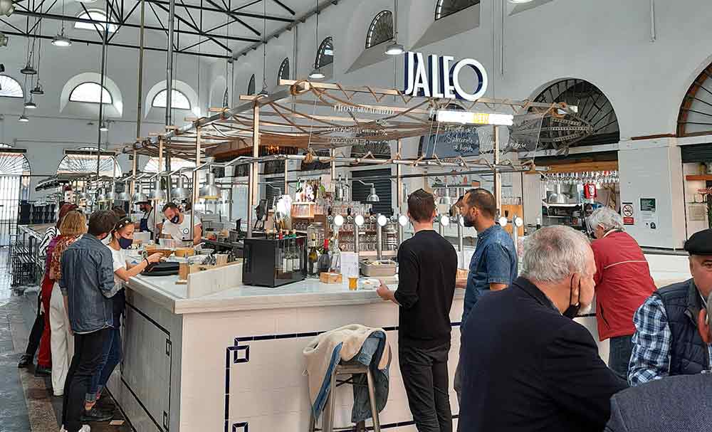 A busy food stall named 'JALEO' inside a modern, airy market hall in Seville, featuring a white tiled bar and industrial-style architecture with arched windows. Patrons stand at the counter enjoying drinks and tapas, highlighting the vibrant social activities in Seville that travellers love.