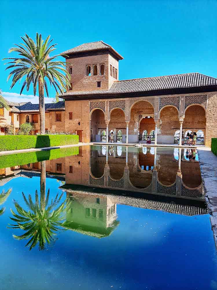The stunning garden at the Alhambra in Granada, Spain showcases its brown hallway, manicured garden and a large pond.