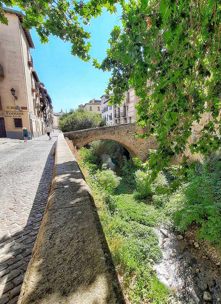 Strolling through the Albaicín District, enjoy the sight of a charming small bridge and sunlit cafes, creating a picturesque scene on a sunny day.