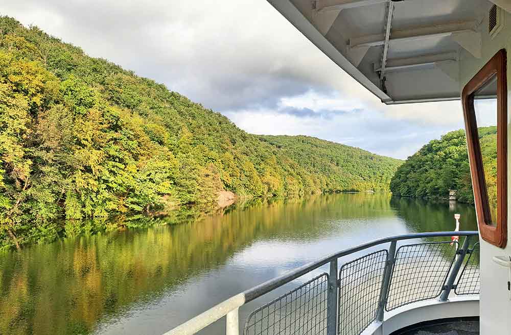 View from the front of a boat looking out over a river. On each side of the river banks there is a forest.  There are greyish clouds in the sky. 