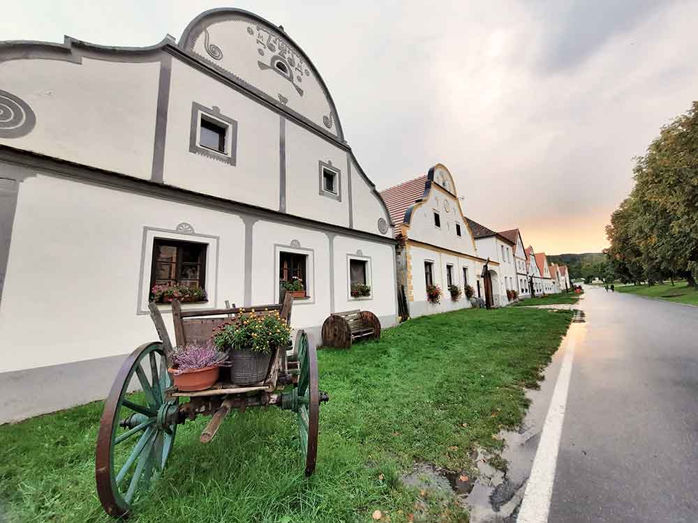 Old farmhouses at Holasovice Medieval Village with a cart and flowers in the foreground. 