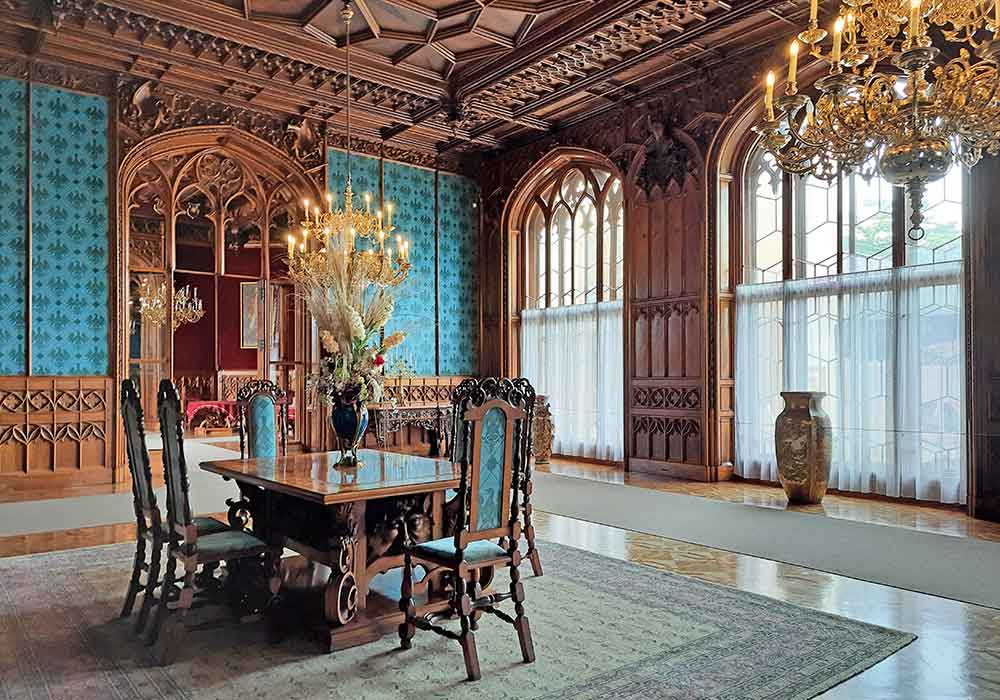 An opulent room at Chateau Lednice with a table in the middle surrounded by six chairs. The walls and ceilings, windows and doors are decorated with detailed wood work, There is blue matching fabric on the chairs and wallpaper.  Two large chandeliers are hanging from the ceiling. 