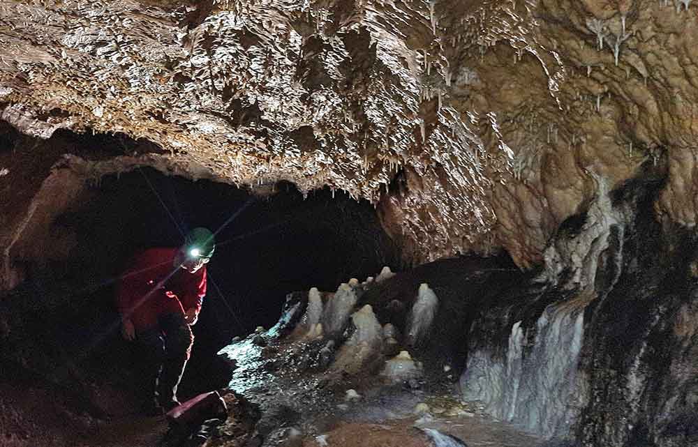 A person in a red overall and green helmet with a flashlight on the front crouching inside a karst