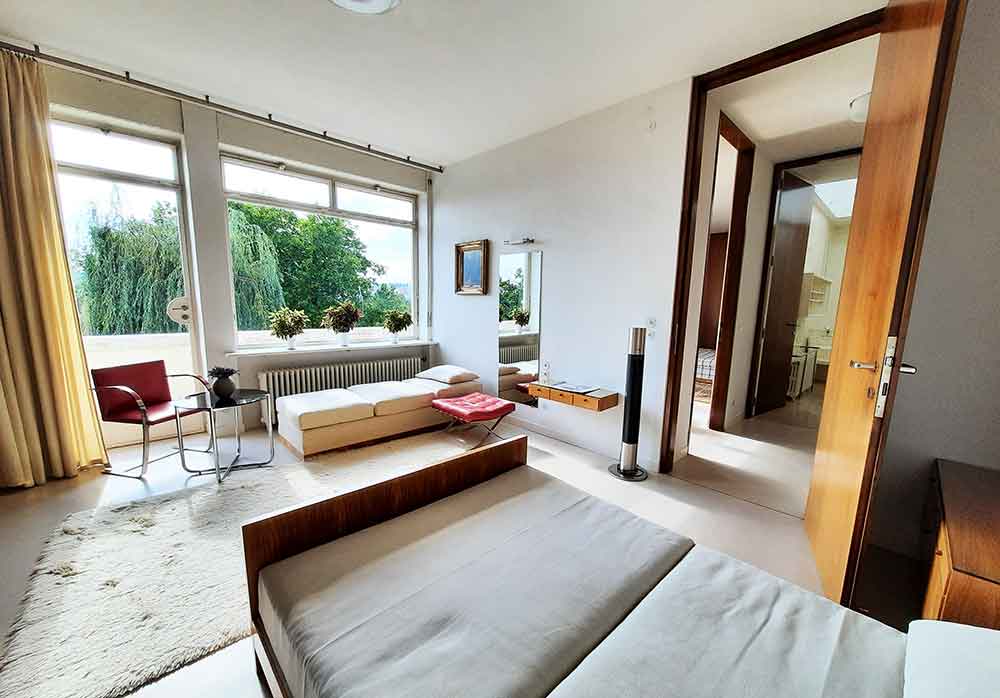 Functionalist-style bedroom at Tugendhat Villa with a large window on the back wall and doors leading through to a hallway. There's a sofa, chair and bed and other furniture in the room. 