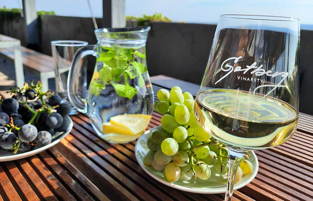 A table laid out with a wine glass with white wine in it, a jug of water with mint and lemon inside and a plate of black grapes and a plate of green grapes. 