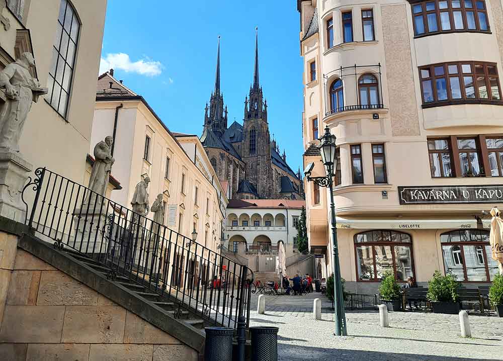 Streets in Brno showing a pair of church spires in the background and with old buildings in the foreground. There are statues in front of the closest building on the left.