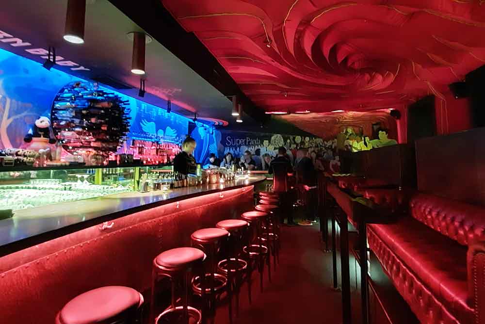 A bar decorated in red velvet with a gigantic red swirl on the ceiling. There are people stood at a table in the back