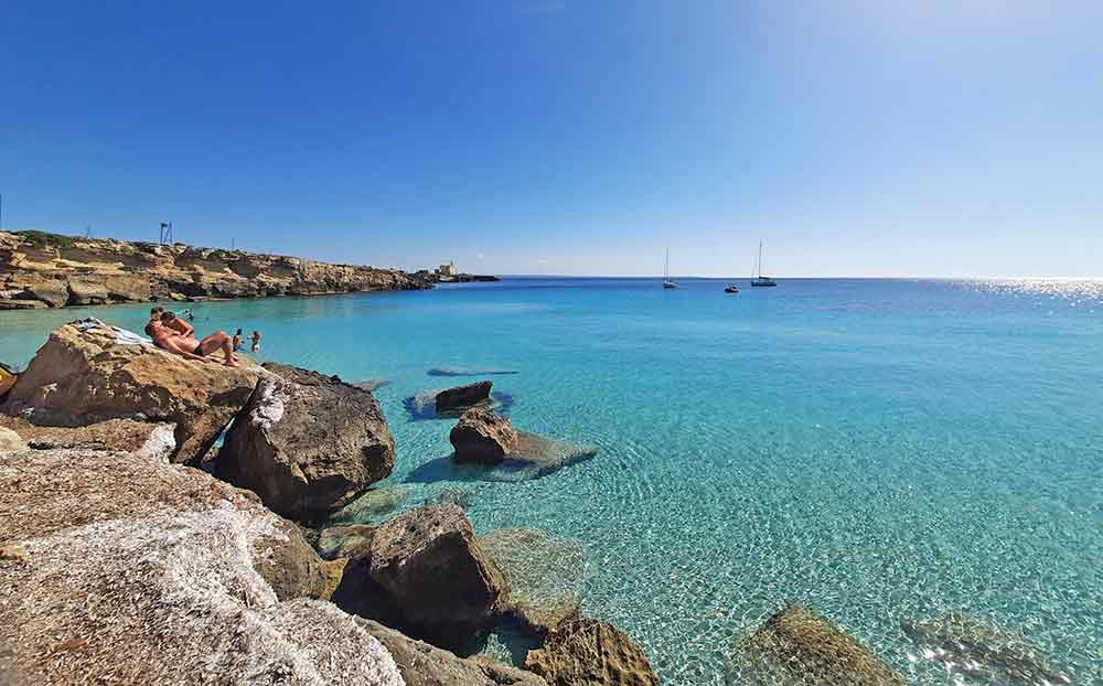 The clear waters of Cala Azzurra on Favignana. This seculuded bay has a small beach and shallow calm water. People are sunbathing on the rocks, and there are a few boats in the distance. 