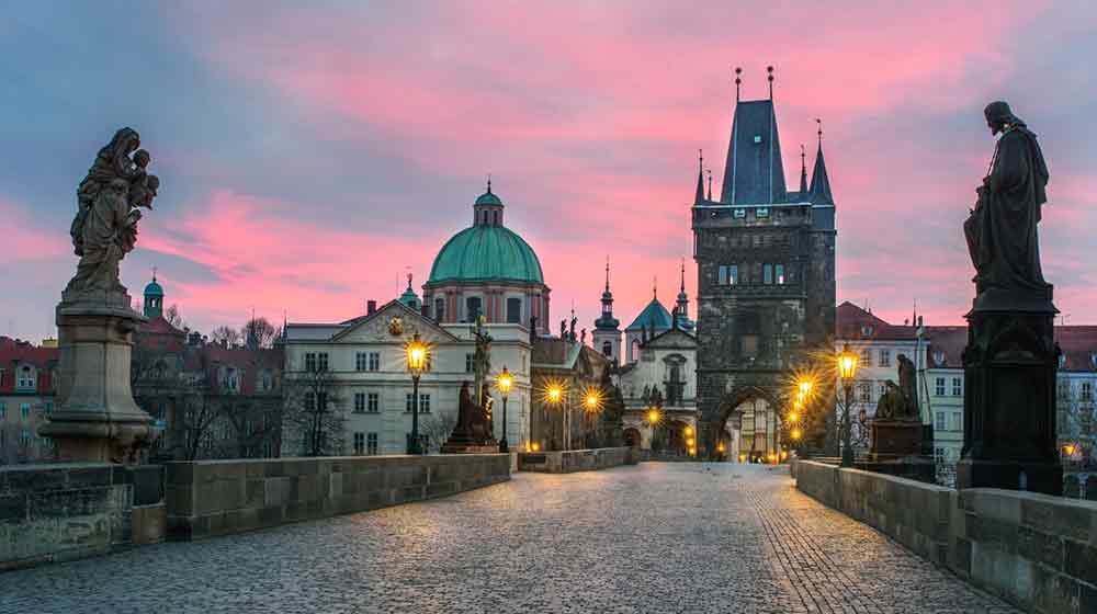 View of Charles Bridge from the bridge looking towards the city. The sky is pink and purple and some streetlights are in the distance. 
