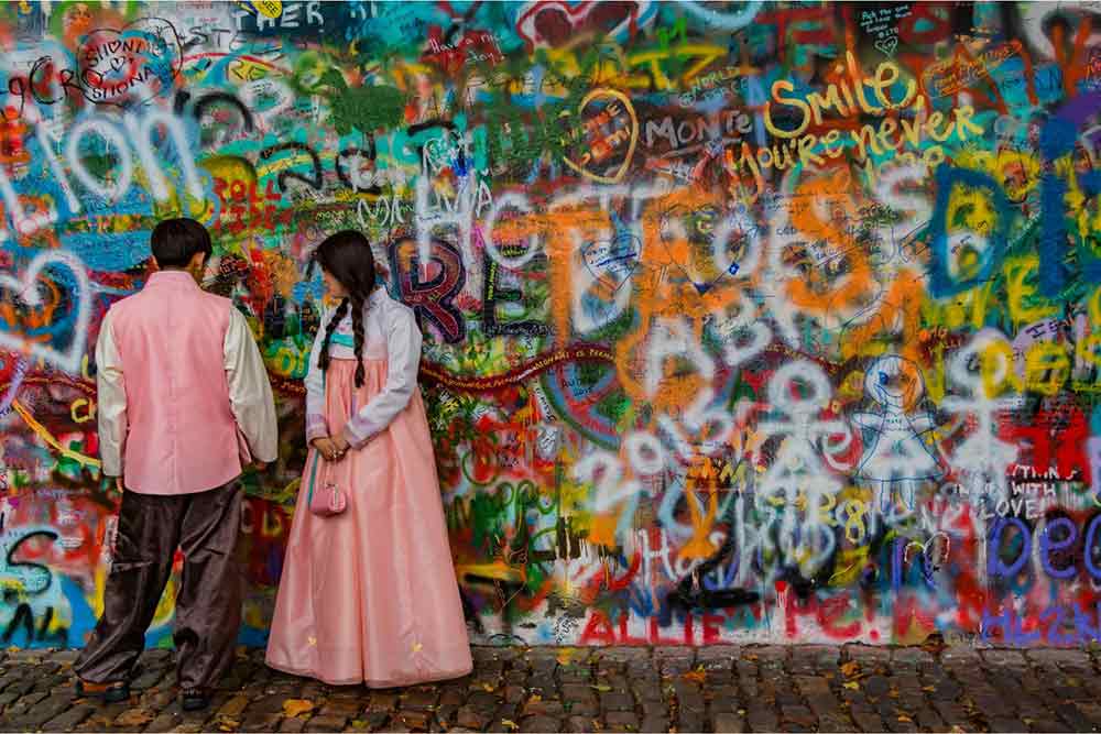 The graffiti covered John Lennon wall in Prague with two people weaing traditional costumes in pink stood in front of it. 