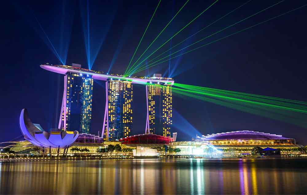 Marina Bay Sands in Singapore featuring a three towers connected by a surfboard-like structure at the top illuminated by colorful lights during the night