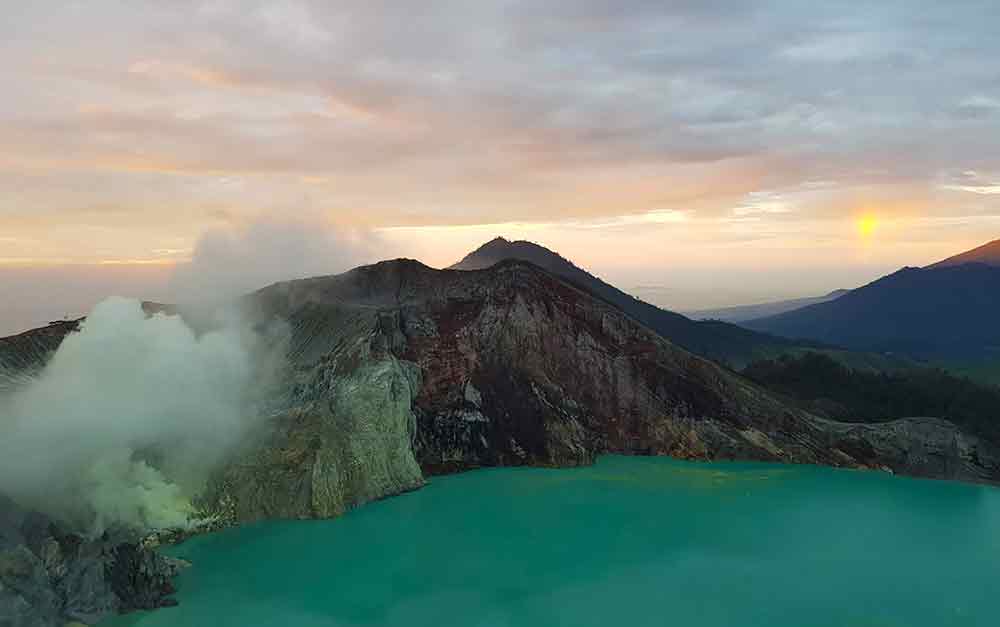 Mt. Ijen, an active volcano, stands alongside the acid lake with white smoke rising from the side during sunset.
