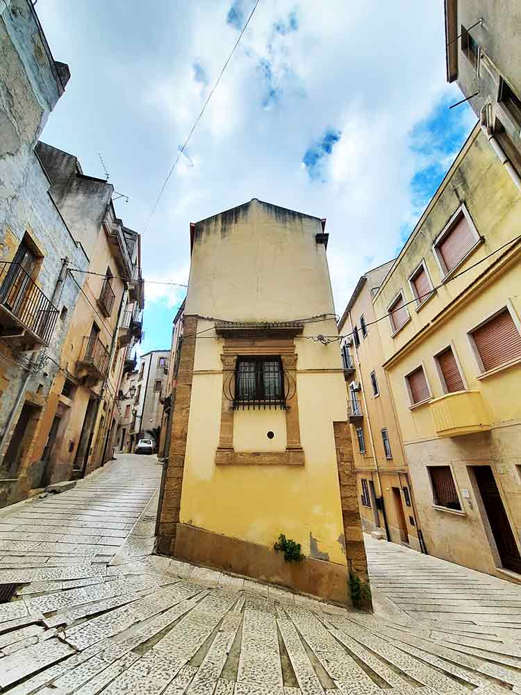 A winding street in Salemi, with a house with a slanted foundation where the road curves around. 