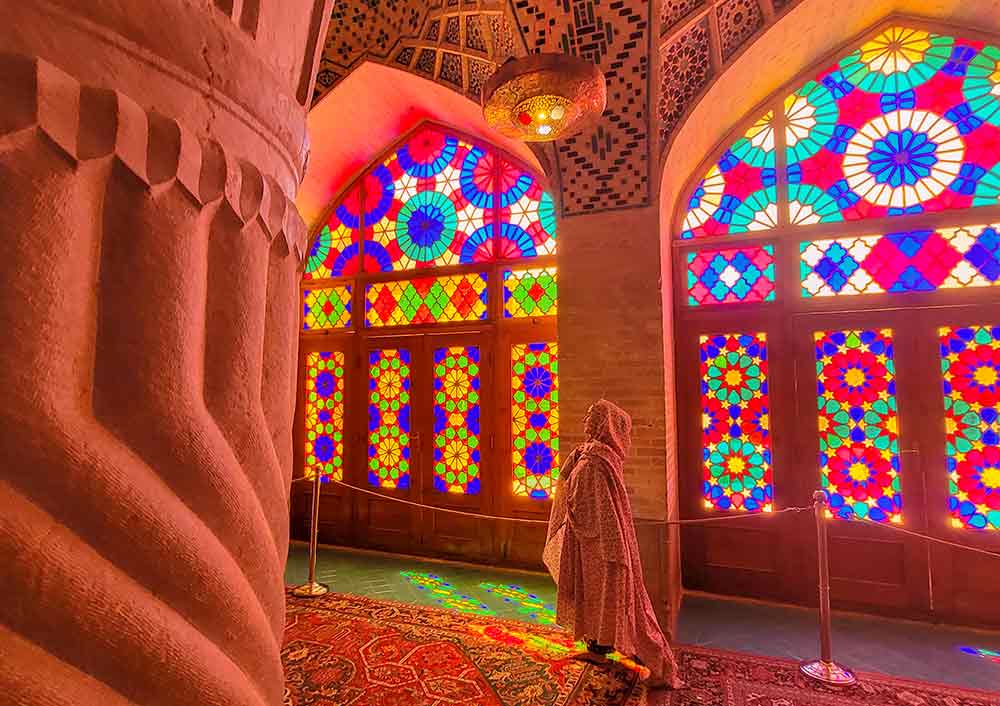 A woman wearing long and covered clothing walks at the Pink Mosque in Iran, featuring pink-tinted structures and colorful mosaic windows.