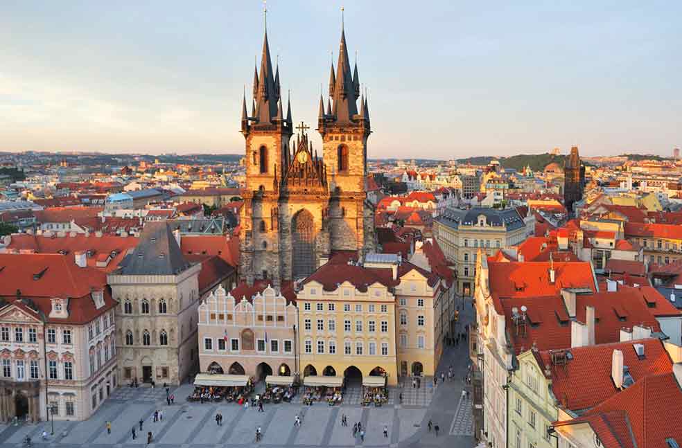 The Old Town Square of Prague with a church with two towers in the background and red rooftops. 