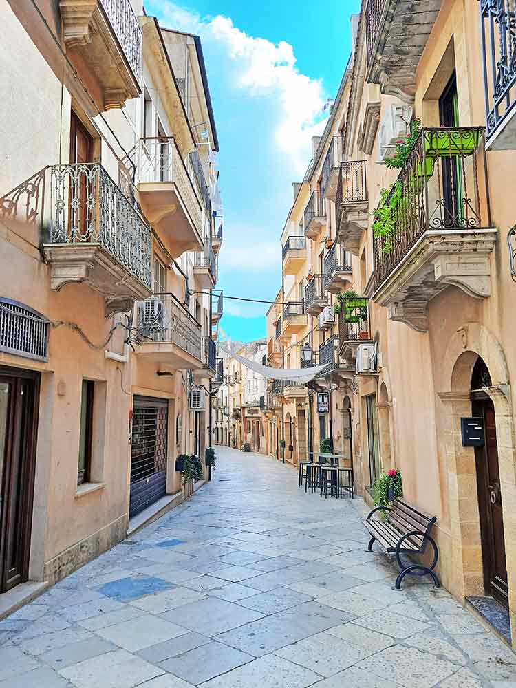 Salemi, Sicily A quiet narrow street in Salemi, Sicily, lined with honey‑coloured stone buildings featuring wooden shutters and arched doorways. Sunlight warms the textured walls and casts gentle shadows, conveying the charm of winter sun in Europe in a small Sicilian town.