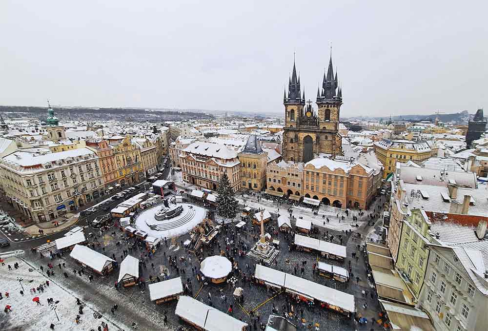 10 Things to Do in Prague at Christmas: A Complete Guide to Christmas in Prague 1 The header image for an article about Prague at Christmas, It shows an aerial image of the old town square in the snow looking down on the Prague Christmas markets. The sky is grey and the buildings are a yellow colour with snow on the roofs around the edge of the square.