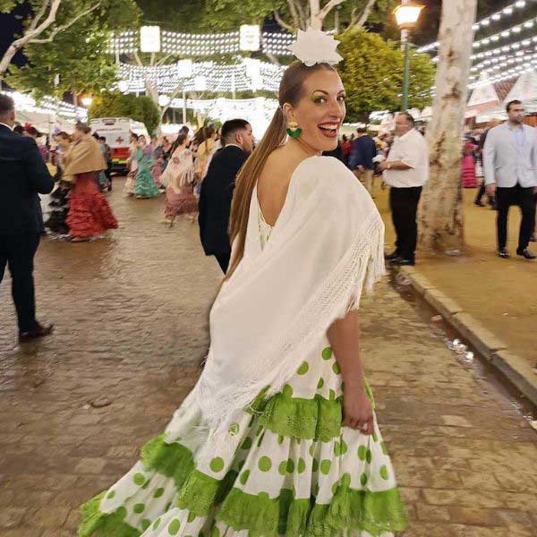 Woman (me) wearing a traditional green and white flamenco dress with a white shawl and flower in her hair, smiling as she walks through a brightly lit fairground at night. The festive atmosphere captures the energy and color of cultural celebrations in southern Spain.