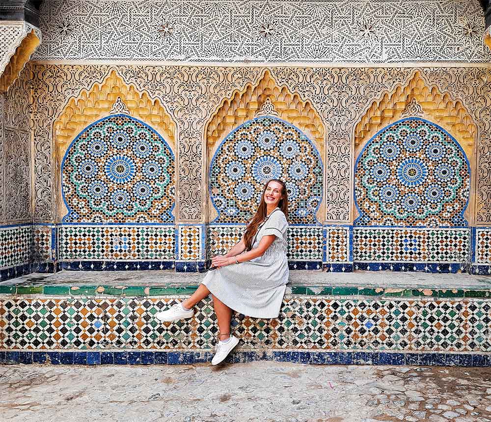 A woman (me) wearing a gray-white dress sits at the center of the stunning mosaic walls in Marrakech