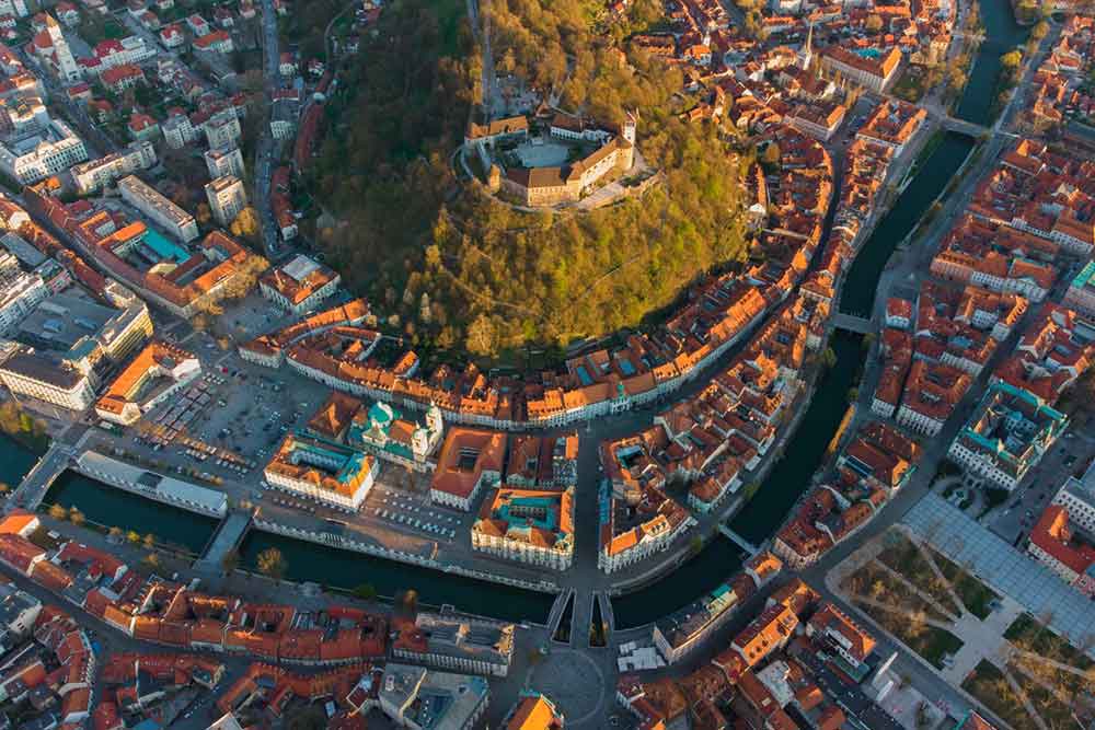 Ljubljana from above features numerous red-roofed houses, buildings, and trees during sunrise.