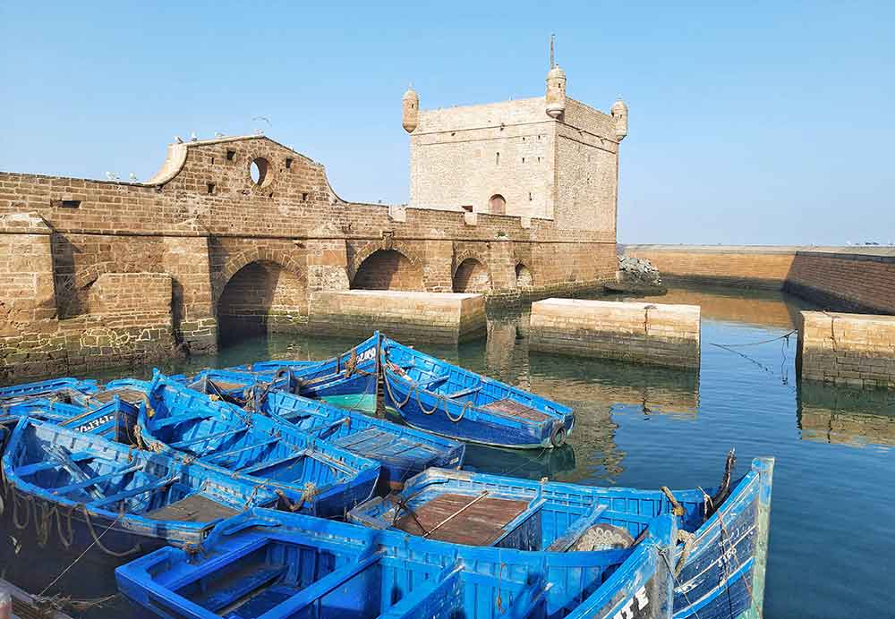 Boats in a harbour at Essaouira old port. There are about 10 little blue rowing boats tied up, there are no people. They are in a shallow hrbour surrounded by the old harbour walls.  self-guided 10 days Morocco itinerary from Casablanca to Marrakech