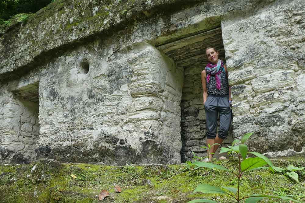 In Plaza de Los Siete Templos, Tikal, a woman stands beside a gray stone covered with moss at the top.
